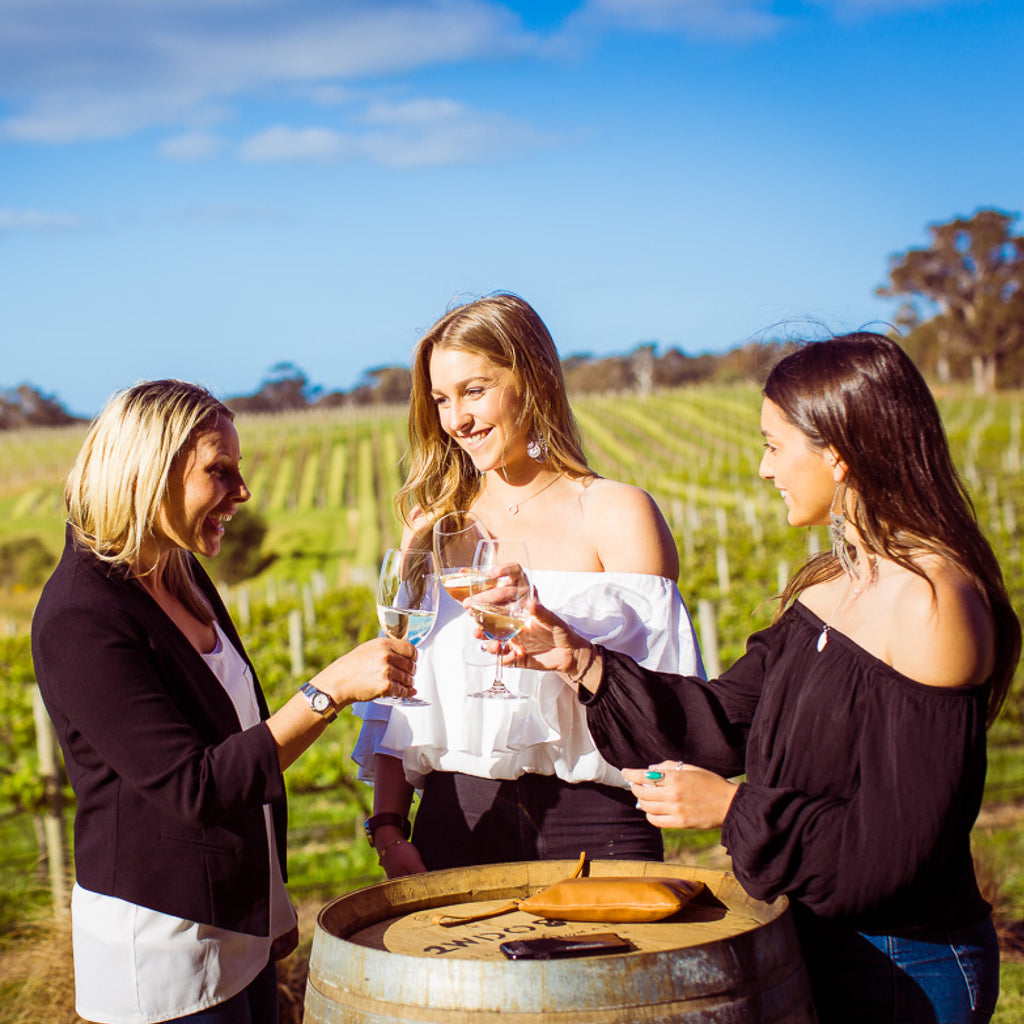 Three women raise their glasses of white wine in a toast on Wills Domain's deck with the vineyard in the background - the perfect setting for a leisurely wine tasting and platters shared with friends.