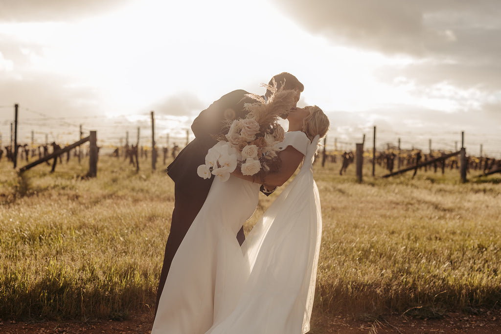 A newly wed couple kiss as the sun sets over Wills Domain vineyards, drenching the image in golden light.