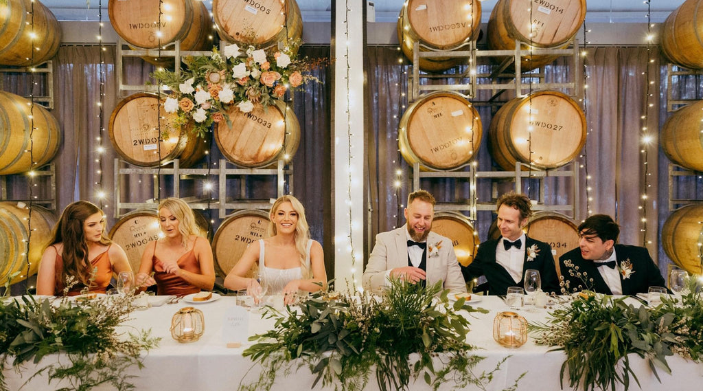 An image of the bridal table with the bride and the groom, bridesmaids and groomsmen, with wine barrels in the background, at Wills Domain's function venue.