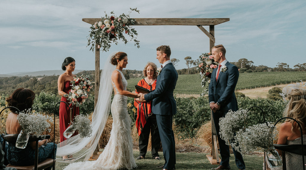 An image of a glamorous bride and groom during their outdoor wedding ceremony, flanked by the best man and the bridesmaid with Wills Domain vineyards in the backdrop.